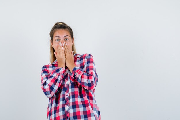 Portrait of young lady keeping hands on mouth in checked shirt and looking happy front view