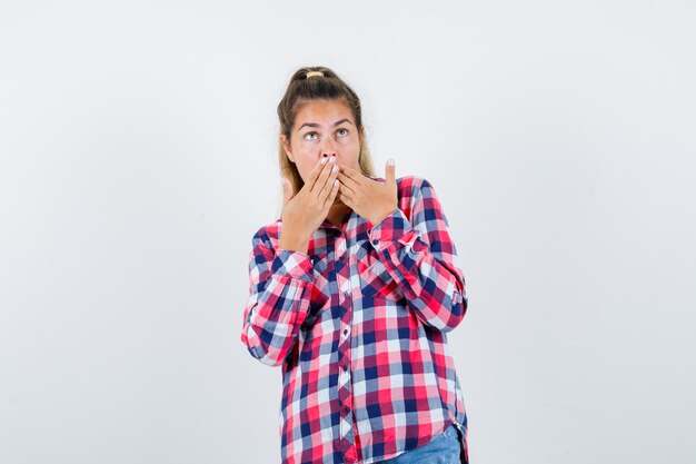 Portrait of young lady keeping hands on mouth in checked shirt and looking scared front view