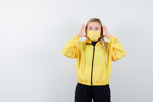 Portrait of young lady keeping raised hands near head in tracksuit, mask and looking irritated front view