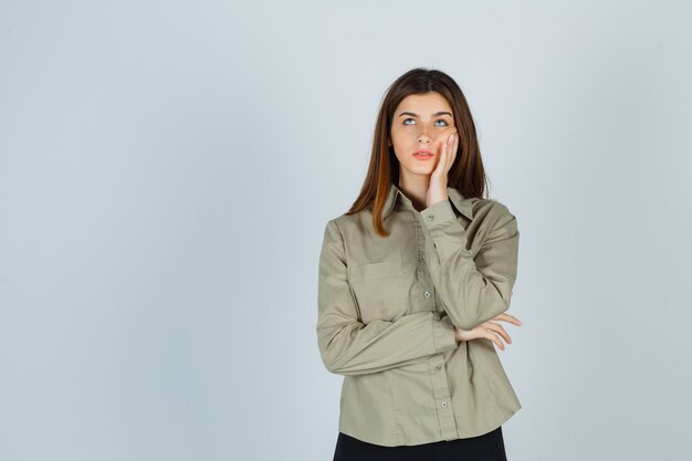 Portrait of young lady leaning cheek on hand while looking up in shirt, skirt and looking thoughtful front view