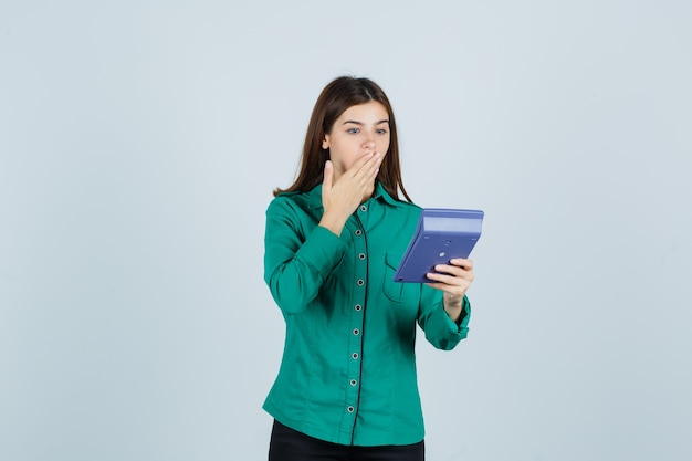 Портрет молодой леди, смотрящей на калькулятор, держась за рот в зеленой рубашке и шокированной взглядом спереди