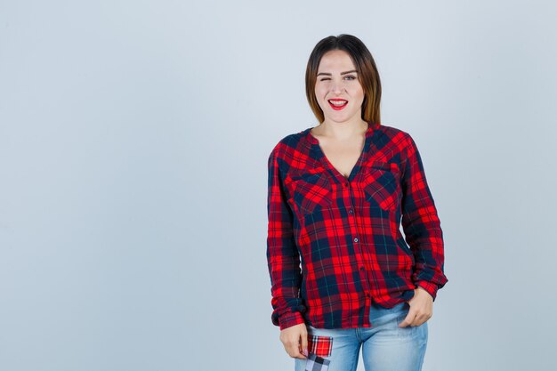 Portrait of young lady looking at front while blinking in checked shirt and looking merry front view