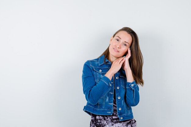 Portrait of young lady pillowing face on her hands in blouse, denim jacket and looking cute front view