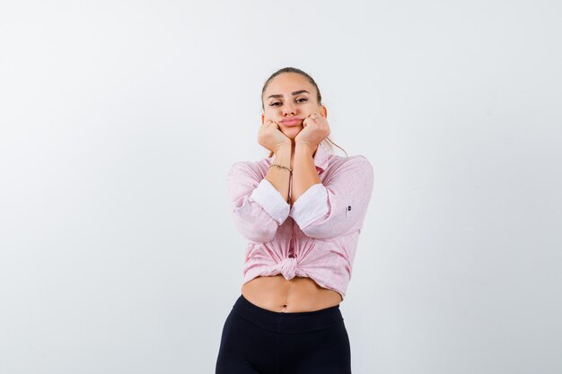 Portrait of young lady pillowing face on her hands in shirt, pants and looking sad front view