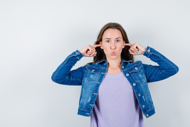 Portrait of young lady plugging ears with fingers, pouting lips in t-shirt, jacket and looking focused front view