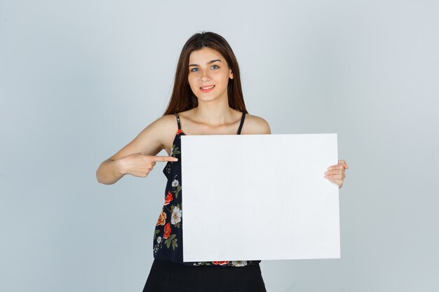 Portrait of young lady pointing at blank canvas in blouse, skirt and looking cheerful front view