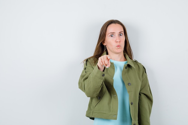 Portrait of young lady pointing at camera in t-shirt, jacket and looking puzzled front view