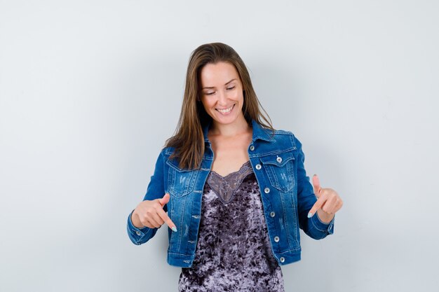 Portrait of young lady pointing down in blouse, denim jacket and looking joyful front view