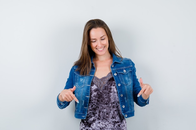 Portrait of young lady pointing down in blouse, denim jacket and looking joyful front view