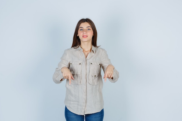Портрет молодой леди, указывая вниз в повседневной одежде, джинсах и оптимистично выглядящей спереди