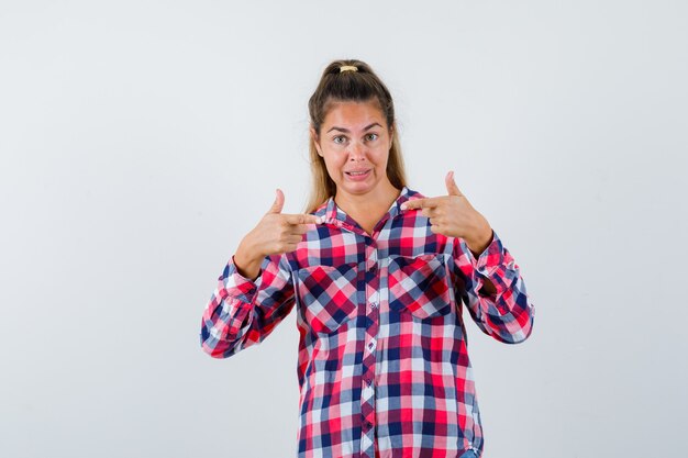 Portrait of young lady pointing at herself in checked shirt and looking puzzled front view