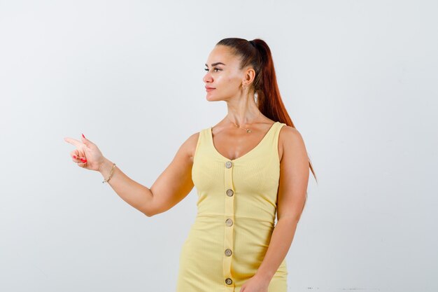 Portrait of young lady pointing left, looking aside in yellow dress and looking focused front view