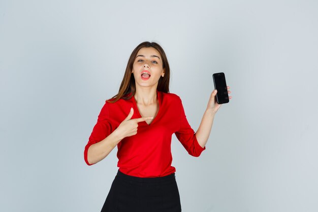 Портрет молодой леди, указывая на мобильный телефон в красной блузке