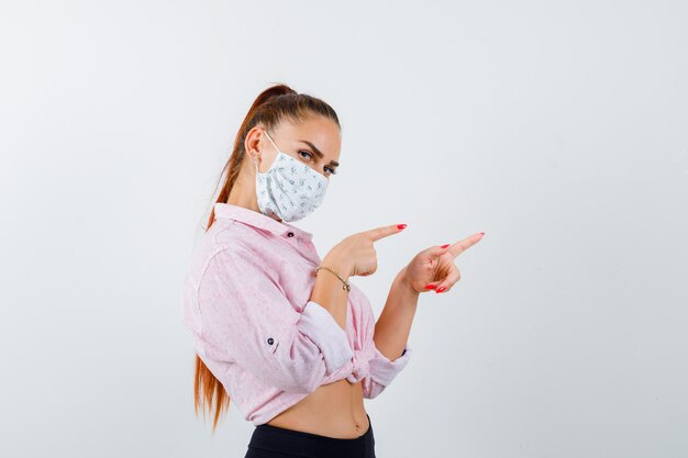 Portrait of young lady pointing right in shirt, mask and looking confident front view