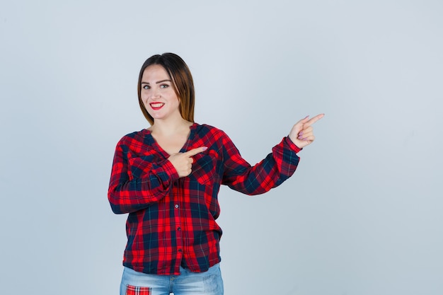 Портрет молодой женщины, указывающей на правую сторону, улыбаясь в клетчатой рубашке, джинсах и веселый вид спереди