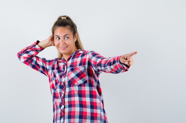 Portrait of young lady pointing at something away in checked shirt and looking joyful front view