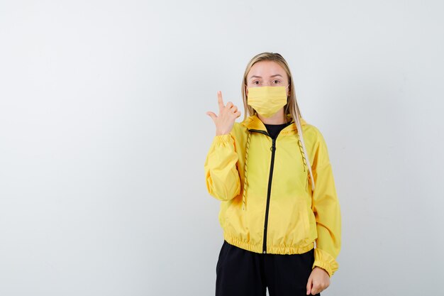 Portrait of young lady pointing up in tracksuit, mask and looking puzzled front view