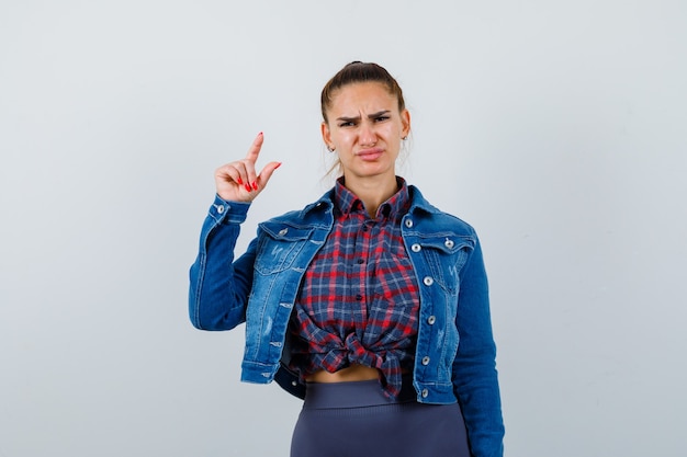 Portrait of young lady pointing up while frowning face in shirt, jacket and looking displeased front view