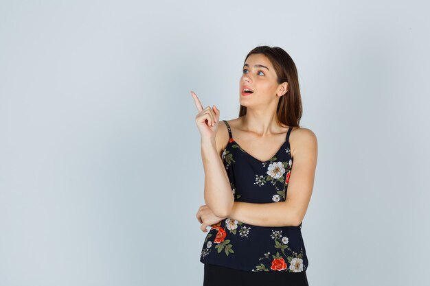 Portrait of young lady pointing at upper left corner in blouse and looking dreamy front view