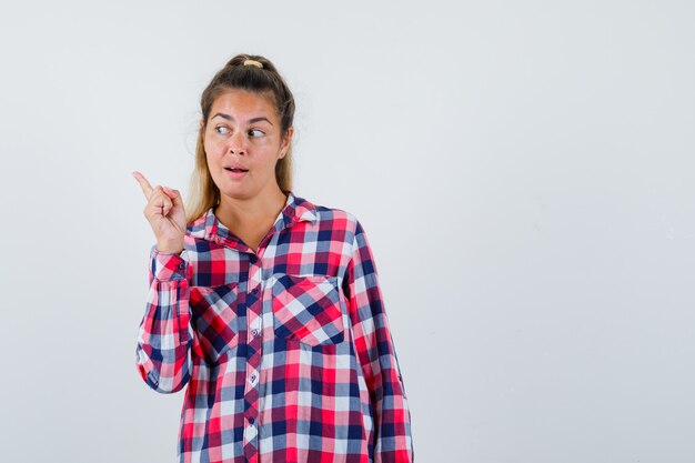 Portrait of young lady pointing at upper left corner in checked shirt and looking curious front view