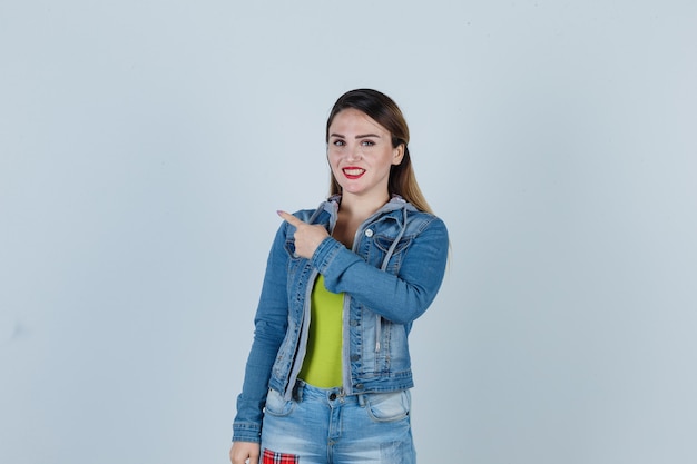 Портрет молодой леди, указывающей на верхний левый угол в джинсовой одежде и веселой смотрящей спереди