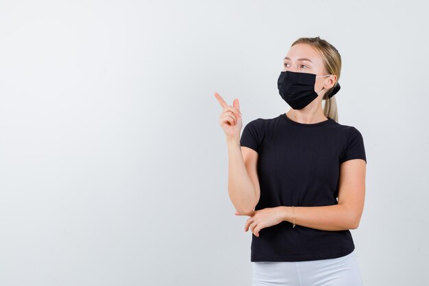 Portrait of young lady pointing at upper left corner in t-shirt, pants, medical mask