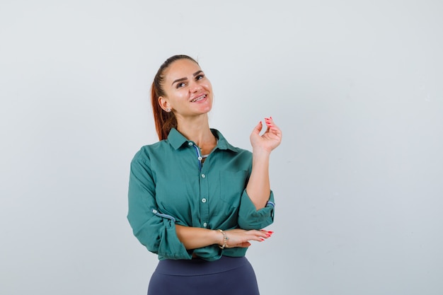 Foto gratuita ritratto di giovane donna in posa con la mano alzata, alzando lo sguardo in camicia verde e guardando una vista frontale sognante