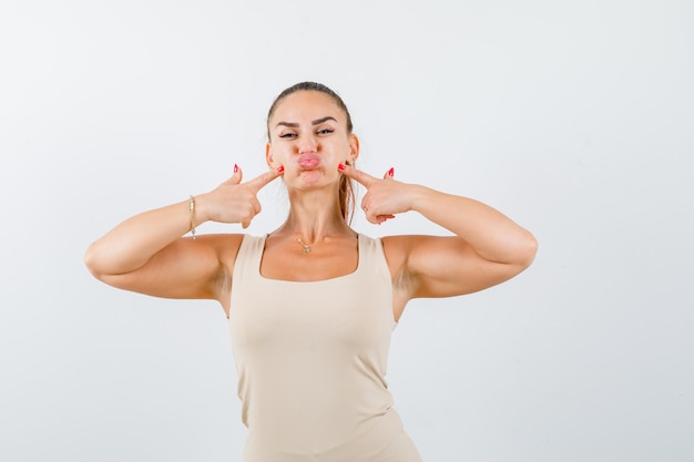 Portrait of young lady pressing fingers on puffy cheeks in tank top and looking funny front view