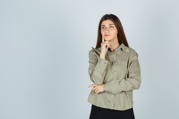 Portrait of young lady propping chin on hand, holding finger on cheek in shirt, skirt and looking pensive front view