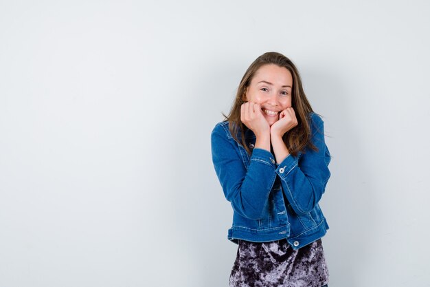 Portrait of young lady propping chin on hands in blouse, denim jacket and looking happy front view