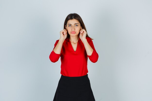 Portrait of young lady pulling earlobes in red blouse, skirt and looking offended