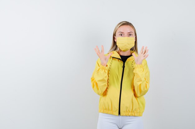 Portrait of young lady raising hands to defend herself in jacket, pants, mask and looking frightened front view