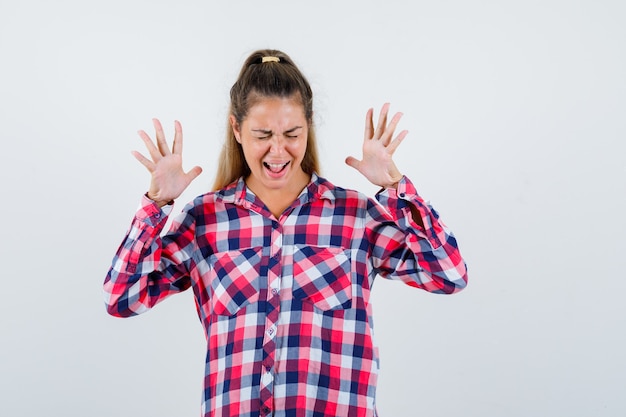 Free photo portrait of young lady raising hands while shouting in checked shirt and looking happy front view