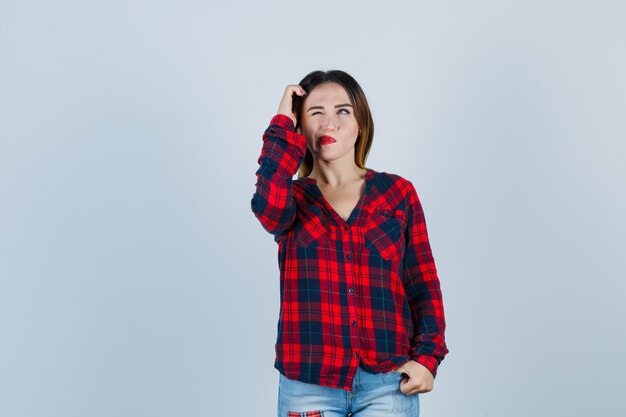 Portrait of young lady scratching head, looking up in checked shirt and looking pensive front view