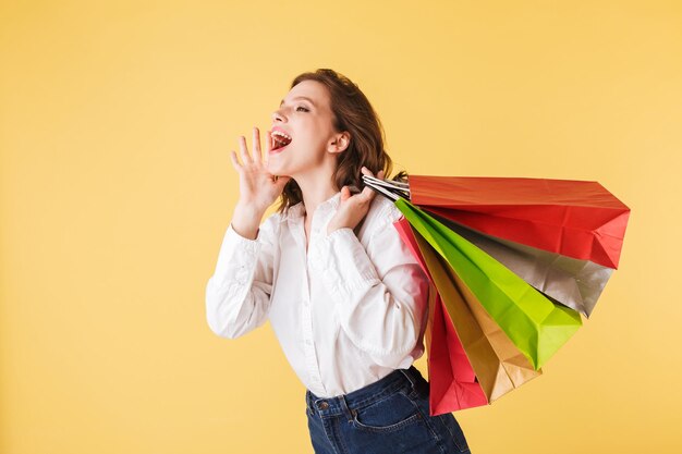 Portrait of young lady in shirt and denim shorts standing with colorful shopping bags in hands and screaming while happily looking aside on over pink background