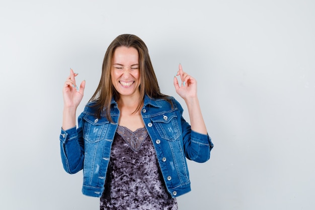 Portrait of young lady showing crossed fingers in blouse, denim jacket and looking cheerful front view