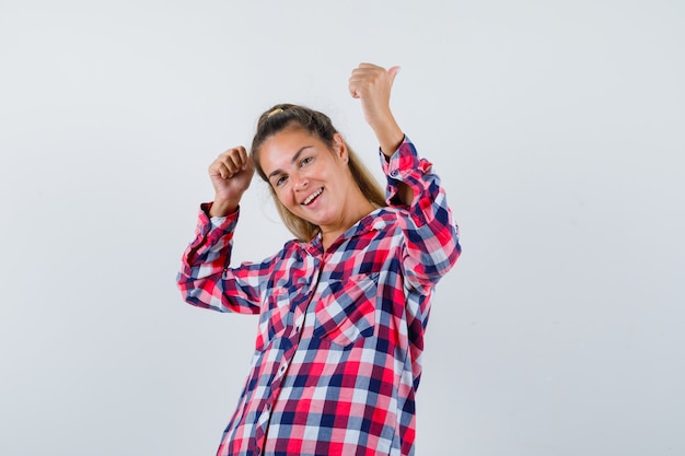 Portrait of young lady showing double thumbs up in checked shirt and looking happy front view