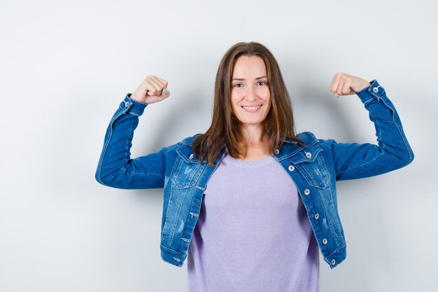 Portrait of young lady showing muscles of arms in t-shirt, jacket and looking merry front view