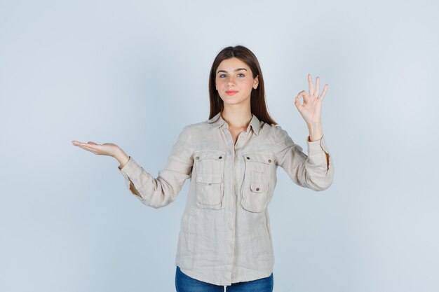 Portrait of young lady showing ok gesture in casual, jeans and looking glad front view