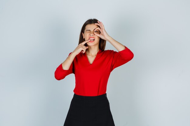 Portrait of young lady showing ok gesture while biting finger in red blouse