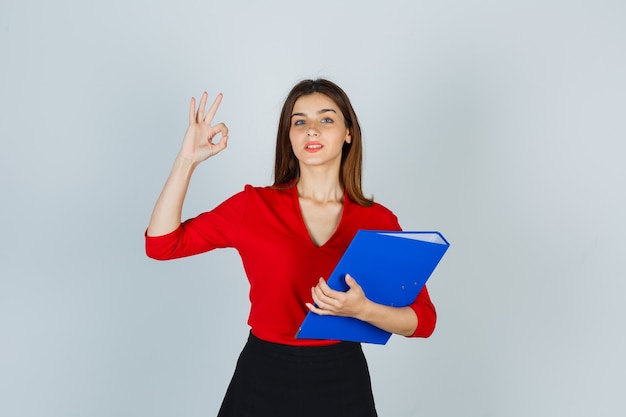 Portrait of young lady showing ok gesture while holding folder in red blouse