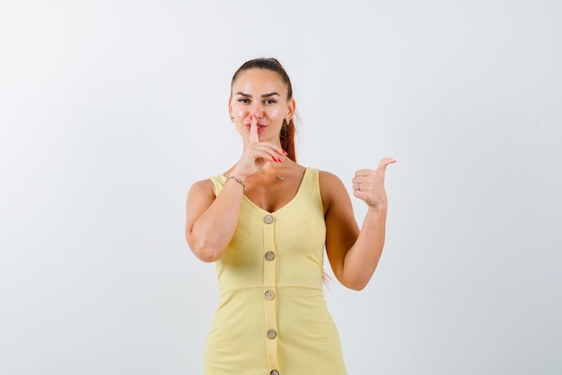 Portrait of young lady showing silence gesture, pointing aside with thumb in yellow dress and looking sensible front view