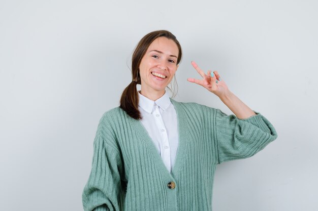 Portrait of young lady showing V-sign in blouse, cardigan and looking joyful front view