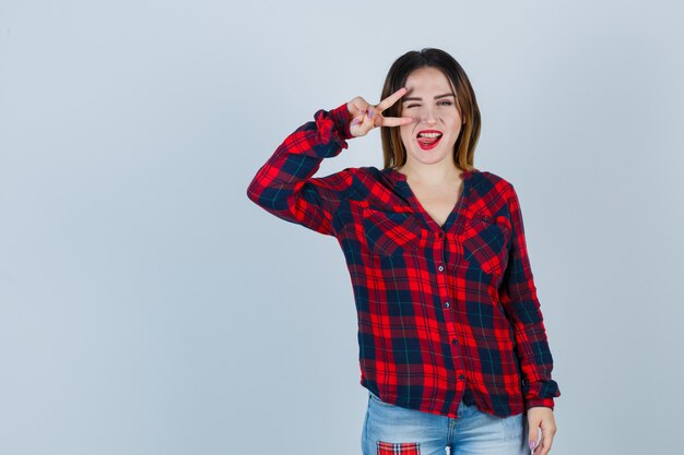 Portrait of young lady showing v-sign near eye, blinking, sticking tongue out in checked shirt and looking lucky front view