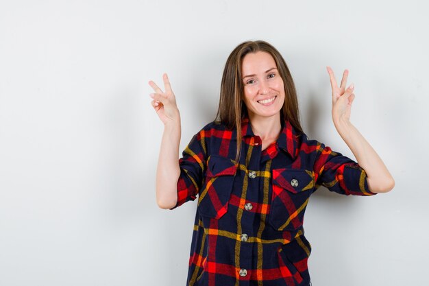 Portrait of young lady showing victory gesture in casual shirt and looking confident front view