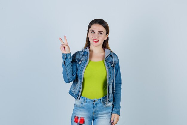 Portrait of young lady showing victory gesture in denim outfit and looking joyful front view