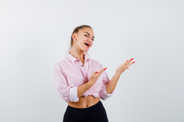 Portrait of young lady showing welcoming gesture in shirt, pants and looking happy front view