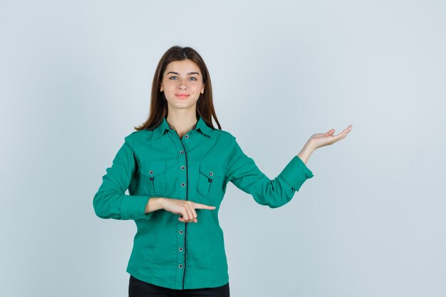 Portrait of young lady showing welcoming gesture while pointing aside in green shirt and looking cheerful front view