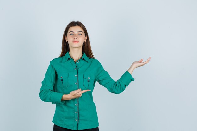 Portrait of young lady showing welcoming gesture while pointing aside in green shirt and looking cheerful front view