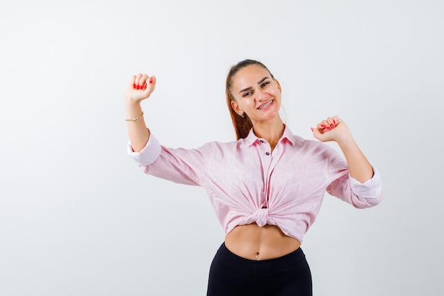 Portrait of young lady showing winner gesture in shirt, pants and looking happy front view